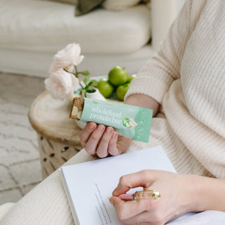 A person holds an apple pie protein bar while journaling on a cozy couch, surrounded by fresh apples and cinnamon sticks, creating a warm, relaxing atmosphere for mindful snacking and reflection