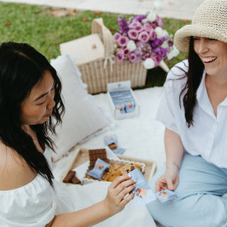 Two friends enjoying a relaxed picnic, connecting over Sneaky Wholefoods Peanut Butter Cookie Nourish Bites, a delicious, plant-based, gluten-free snack